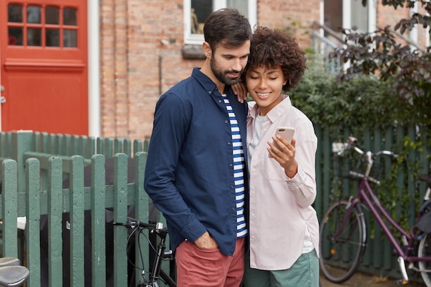 Horizontal shot of cheerful mixed race young woman and man read information on mobile phone