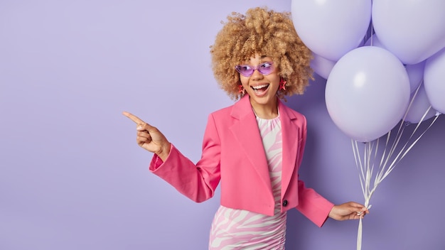 Free photo horizontal shot of cheerful woman dressed in fashionable clothes holds bunch of inflated balloons celebrates university leaving organizes party points away on copy space isolated on purple background