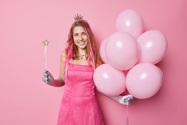 Горизонтальный снимок веселой женщины готовится к празднованию, носит праздничное платье, длинные перчатки, корона держит волшебную палочку и кучу надутых воздушных шаров, изолированных на розовом фоне Концепция праздников