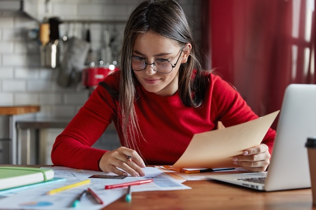 Horizontal shot of cheerful woman sits at kitchen table reviews finances, sits in front of opened laptop