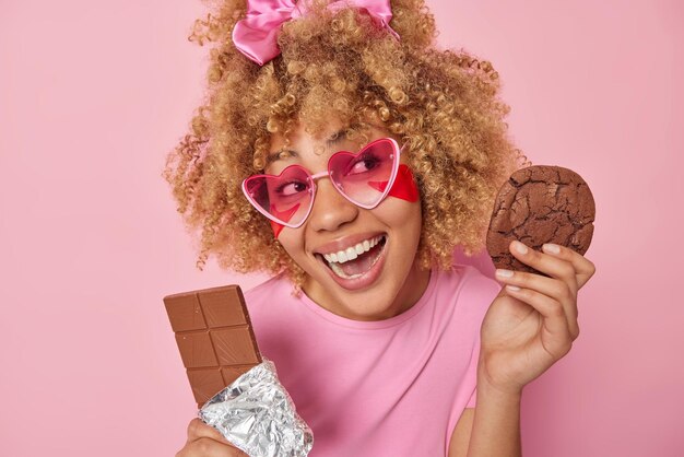 Horizontal shot of cheerful woman with curly hair holds bar of chocolate and delicious cookie has sweet tooth enjoys eating unhealthy harmful food applies beauty patches under eyes poses indoor