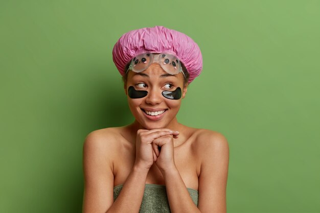 Horizontal shot of cheerful young Afro American woman keeps hands under chin smiles gently applies beauty pads under eyes wrapped in bath towel isolated over green wall