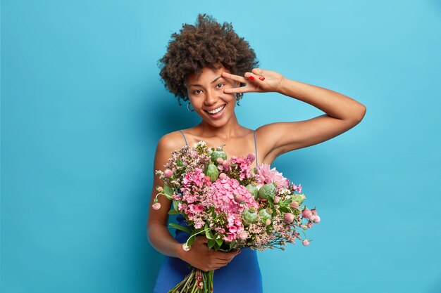 쾌활한 젊은 여자의 가로 샷은 평화 제스처가 꽃 미소의 큰 꽃다발을 보유하고 광범위하게 파란색 벽에 고립 된 축제 분위기를 보여줍니다