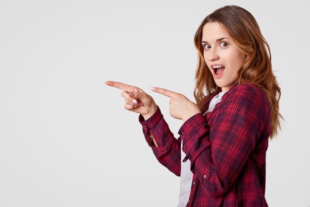 Horizontal shot of cheerful young woman with long hair, points with both fore fingers at empty space, wears casual checkered shirt