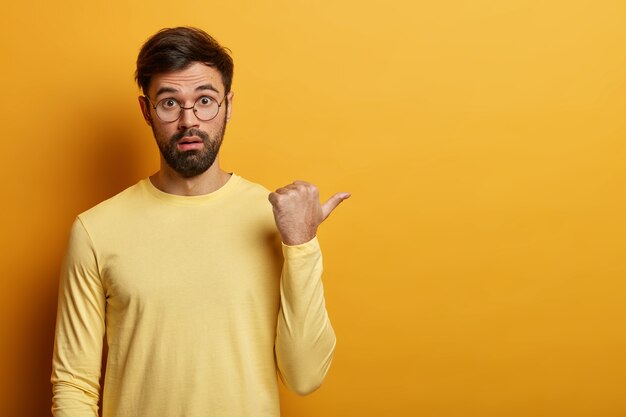 Horizontal shot of confused bearded man points thumb right, discusses discount offer, tells about amazing unbelievable sale, wears glasses and sweater, copy space for your promotional content