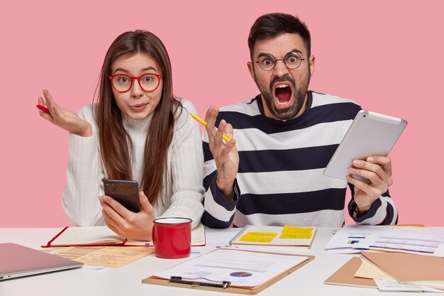 Horizontal shot of confused female and annoyed man discuss graphs, work on university project, use modern technologies, do homework together, isolated over pink wall. Youth and work concept