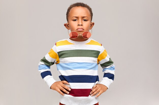 Horizontal shot of cool handsome Afro American child having confident facial expression keeping hands on his waist, stylish round pink shades falling down his nose. Childhood and fashion concept