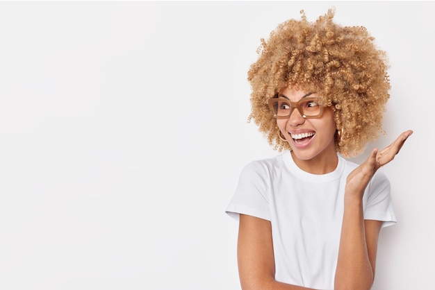 Free photo horizontal shot of crazy curly woman reacts emotionally on something keeps palm raised smiles happily wears casual t shirt and spectacles poses against white background copy space for your promo