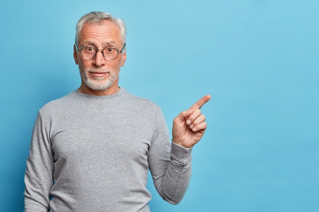 Horizontal shot of curious grey haired senior man points index finger away in blank space shows place for your advertisement wears casual jumper isolated over blue wall
