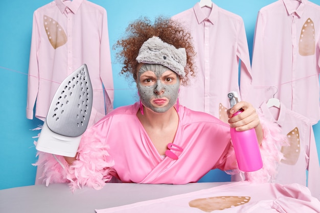Horizontal shot of curly haird young woman looks angrily  holds bottle spray with water and electric iron wears domestic robe sleepmask on forehead poses against ironed clothes around