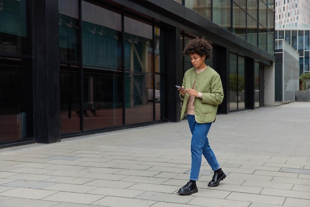 Horizontal shot of curly haired woman enjoys free time in city holds mobile phone chats online walks in urban setting