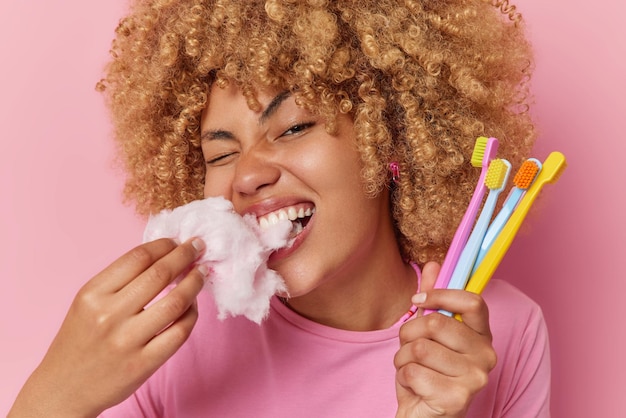 Горизонтальный снимок кудрявой молодой женщины кусает аппетитную сладкую вату, вредную для зубов, держит много зубных щеток в ручных позах на розовом фоне Сладкая нездоровая еда и зависимость от сахара