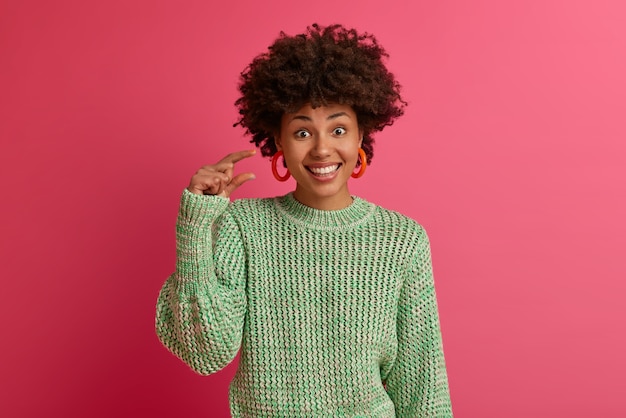 Horizontal shot of dark skinned african american woman shows small amout gesture, measures little item, smiles pleasantly, wears knitted sweater, isolated on pink wall. body language concept