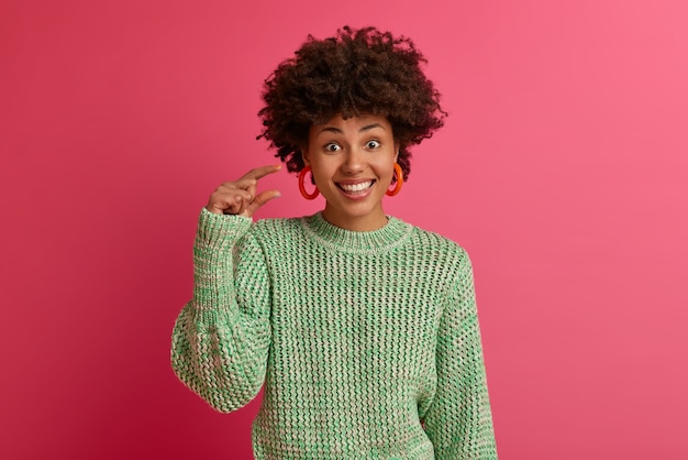Horizontal shot of dark skinned African American woman shows small amout gesture, measures little item, smiles pleasantly, wears knitted sweater, isolated on pink wall. Body language concept