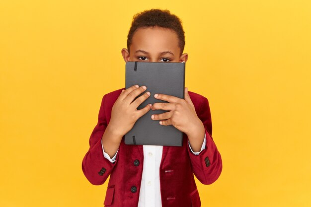 Horizontal shot of dark skinned schoolboy wearing velvet jacket holding black notebook covering face, doing homework.