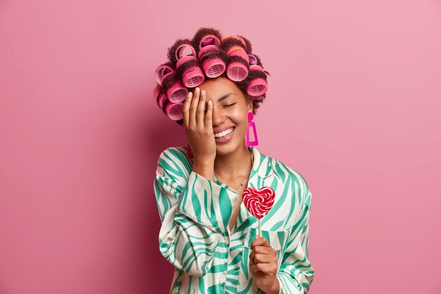 Horizontal shot of dark skinned woman stands with eyes closed, makes face palm, has fun at home, holds delicious heart shaped lollipop, wears hair rollers, smiles broadly, isolated on pink wall