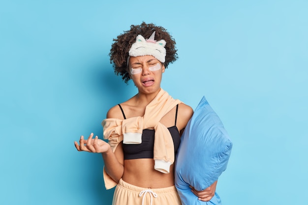 Horizontal shot of dejected Afro American woman cries from despair dressed in nightwear holds pillow suffers insomnia applies collagen patches isolated over blue wall