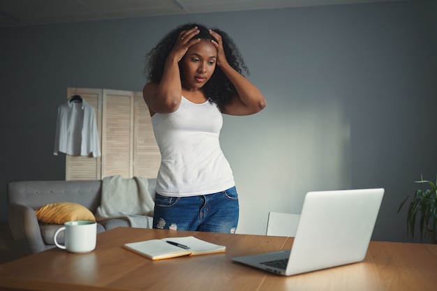 Horizontal shot of desperate young Afro American woman freelancer holding hands on her head, feeling stressed and panic because of deadline or computer problem, standing at desk with open laptop