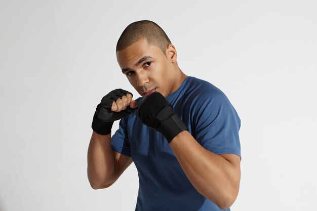 Horizontal shot of determined young Afro American male with atheltic body wearing training gloves posing in defensive position, holding pumped fists in front of his face and staring at camera