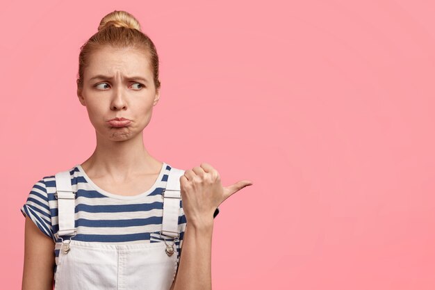 Horizontal shot of displeased gloomy blonde female purses lips and points aside with thumb, feels discontent with something, poses over pink wall