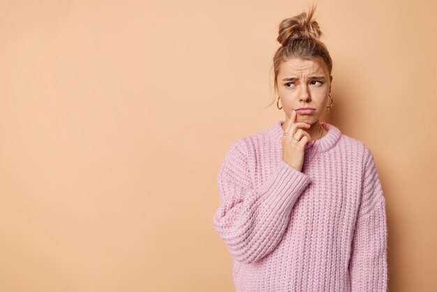 Horizontal shot of displeased sad woman purses lips looks unhappily away wears loose knnitted sweater poses against beige background with copy space on left for your advertisement has bad mood