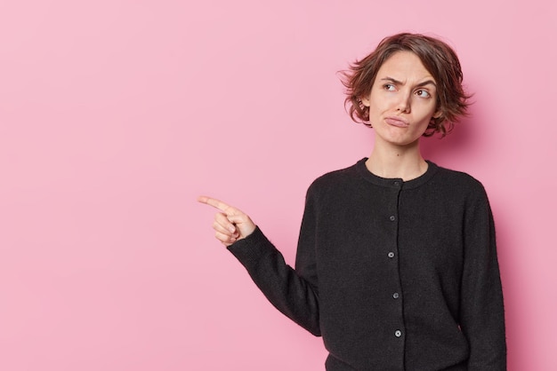 Horizontal shot of displeased woman with short hair raises eyebrows sulks face points index finger away on blank space advertises something isolated over pink background. Look at this promo.