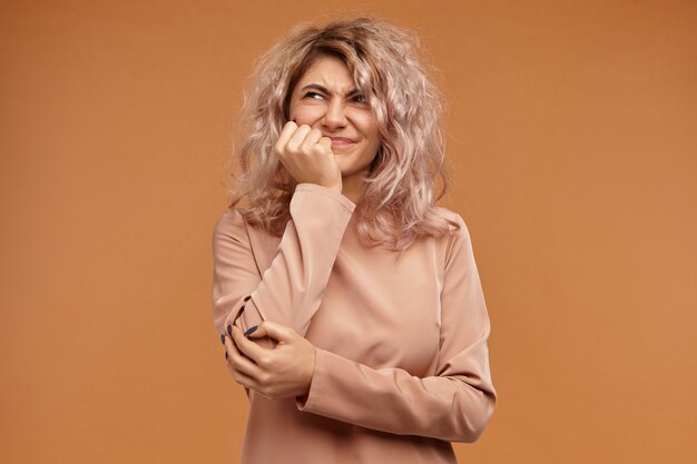 Horizontal shot of displeased young woman with stylish hairdo grimacing holding hand on her cheek, having painful look, suffering form terrible toothache