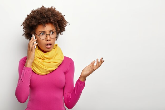 Horizontal shot of dissatisfied dark skinned woman raises palm has puzzled expression, negative conversation via cellphone, dressed in vivid clothes and transparent spectacles