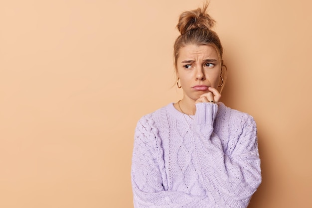 Free photo horizontal shot of dissatisfied frustrated woman looks sadly away purses lips wants to cry being offended has thoughtful expression wears knitted sweater poses against beige studio background