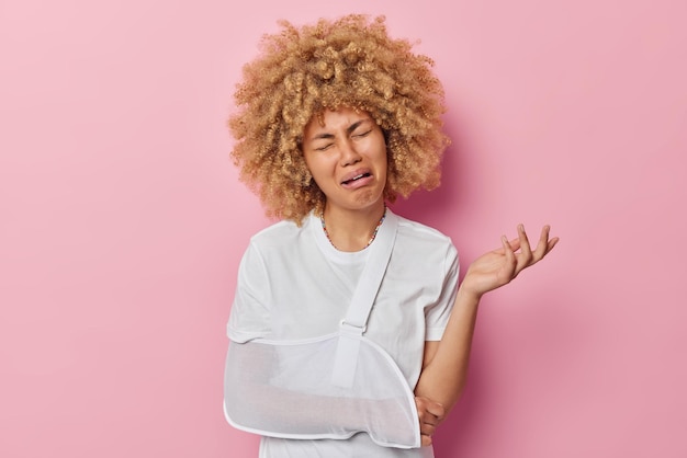 Horizontal shot of dissatisfied young woman cries from despair suffers from pain wears sling on broken arm after visiting orthopedic clinic isolated over pink background Health problems concept