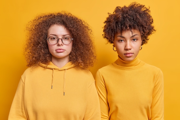 Горизонтальный снимок разных женщин-компаньонов, стоящих рядом друг с другом, выглядящих серьезно, внимательных взглядов, одетых небрежно, слушайте информацию, изолированную над желтой стеной
