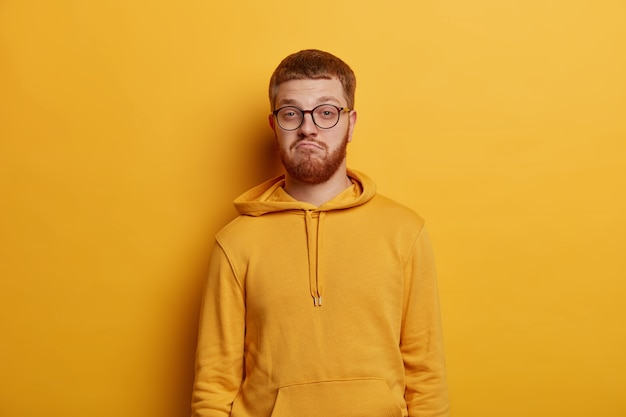 Horizontal shot of doubtful bearded man with ginger hair and beard purses lips and looks confusingly , hears puzzled news, has specific appearance, wears yellow hoodie and spectacles