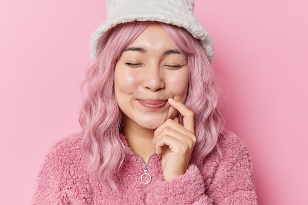 Free photo horizontal shot of dreamy delighted asian woman closes eyes keeps finger near corner of lips has pink dyed hair dressed in fur coat and winter hat isolated over pink background happy feelings