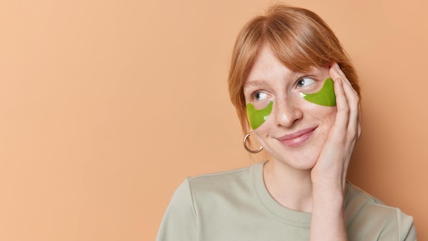 Horizontal shot of dreamy millennial girl with red hair freckled skin applies green hydrogel patches under eyes to remove puffiness isolated over brown background blank copy space for your text