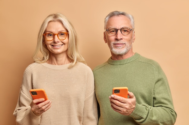 老夫婦の水平方向のショットは、現代の技術を使用してスマートフォンを保持し、ワイヤレスインターネットに接続されたテキストメッセージを読み取ります茶色の壁に隔離されたカジュアルなジャンパーを着用します