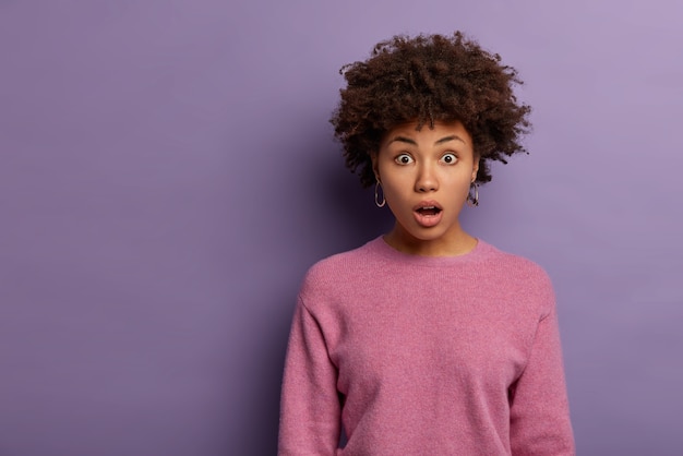 Horizontal shot of embarrassed curly young woman gasps from wonder, opens mouth in wow sound, hears stunning news, wears rosy jumper, found out unexpected rumors, isolated over purple wall