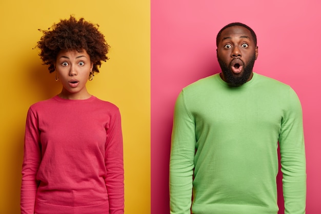 Free photo horizontal shot of embarrassed shocked ethnic couple stare with bugged eyes , fascinated by something terrible, gasps from wonder, wear pink and green jumpers, pose over colorful  wall