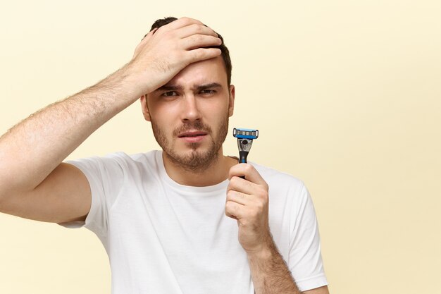 Horizontal shot of emotional scared young Caucasian man holding safety razor and keeping hand on cheek being afraid of shaving.