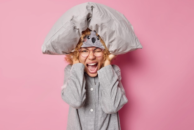 Free photo horizontal shot of emotional woman exclaims loudly cannot fall asleep because of noise keeps soft pillow over head covers ears dressed in nightwear isolated over pink background sleep interruption