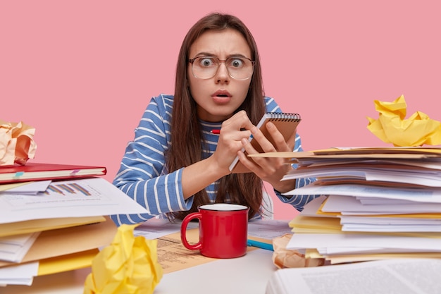 Free photo horizontal shot of emotive caucasian woman writes down notes in spiral notepad, wears optical glasses and striped clothes, enjoys hot drink