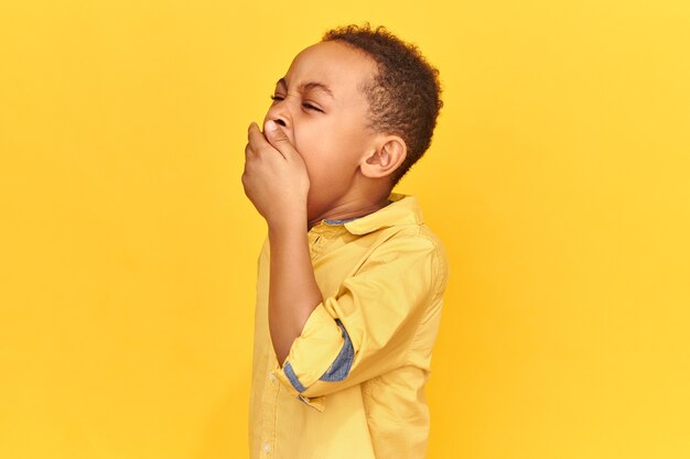 Горизонтальный снимок измученного сонного африканского школьника в желтой рубашке, прикрывающего рот рукой, зевающей, уставшего после долгого утомительного дня. Концепция скуки, сна, времени сна и постельного белья