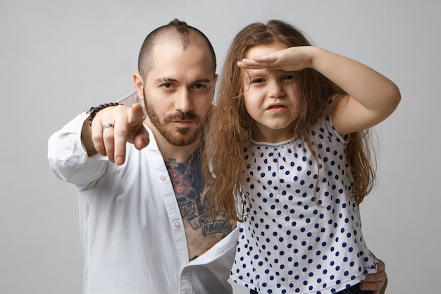 Free photo horizontal shot of fashionable young father with beard and tattoo on chest pointing finger at camera