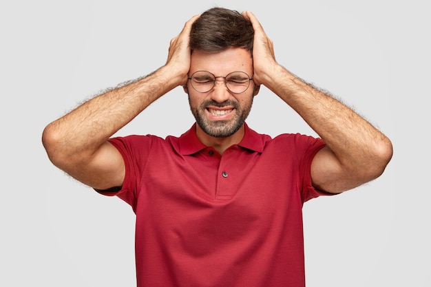 Colpo orizzontale di fatica giovane maschio con la barba lunga euroepea tiene le mani sulla testa, si sente esausto dopo il lavoro in ufficio, ha mal di testa, indossa una maglietta rossa brillante casual, isolato sopra il muro bianco