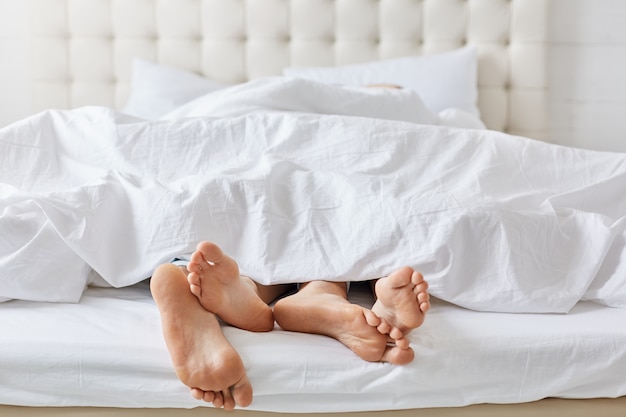 Horizontal shot of feet of couple under white bedclothes in bed at bedroom