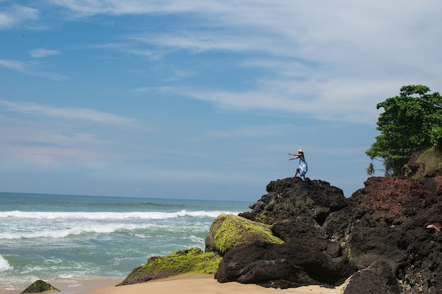 Horizontal shot of a female with a hat and blue dress on a rocky beach