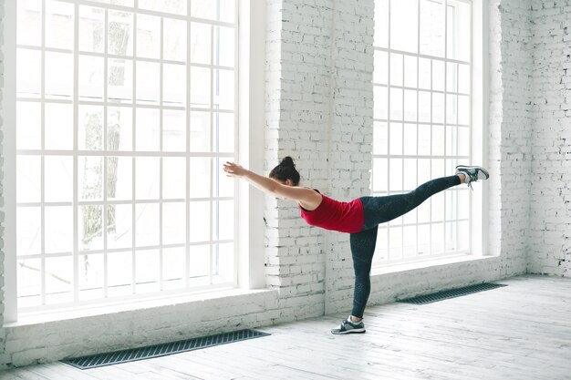 Horizontal shot of fit attractive young woman wearing stylish sports clothes practicing yoga asanas indoors for strengthening thighs, calves, ankles and back, improving balance and concentration