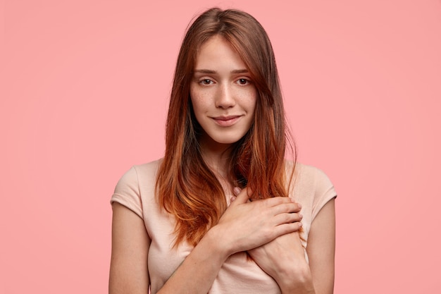 Horizontal shot of friendly-looking female with freckled skin, keeps hands on heart, looks happily at camera