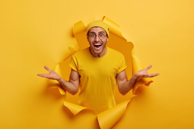 Horizontal shot of friendly looking happy Caucasian man spreads hands sideways, presents something cool, dressed in casual clothes, poses in paper hole alone