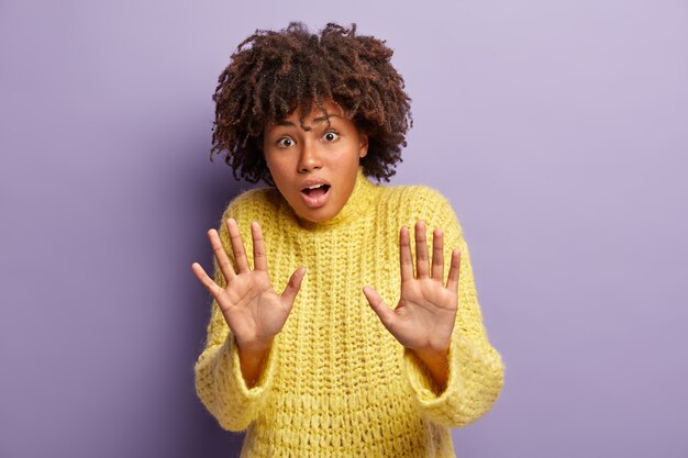 Horizontal shot of frightened scared woman with Afro hairstyle, stretches palms in protective gesture