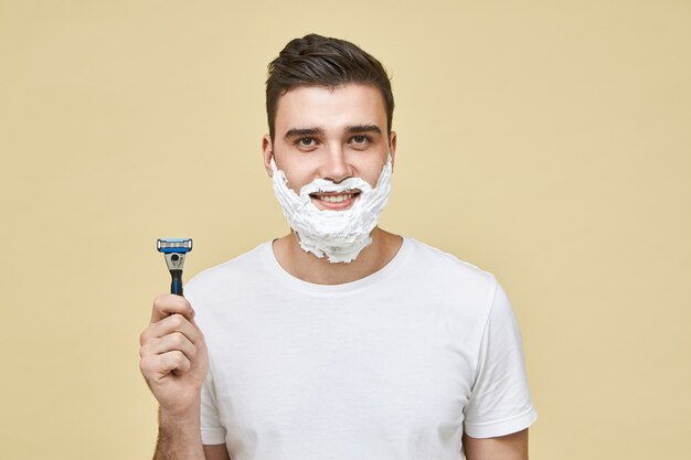 Horizontal shot of funny handsome young male with white foam on his face with smile, holding shaving stick, going ti shave beard, doing morning routine. Grooming and men's beauty